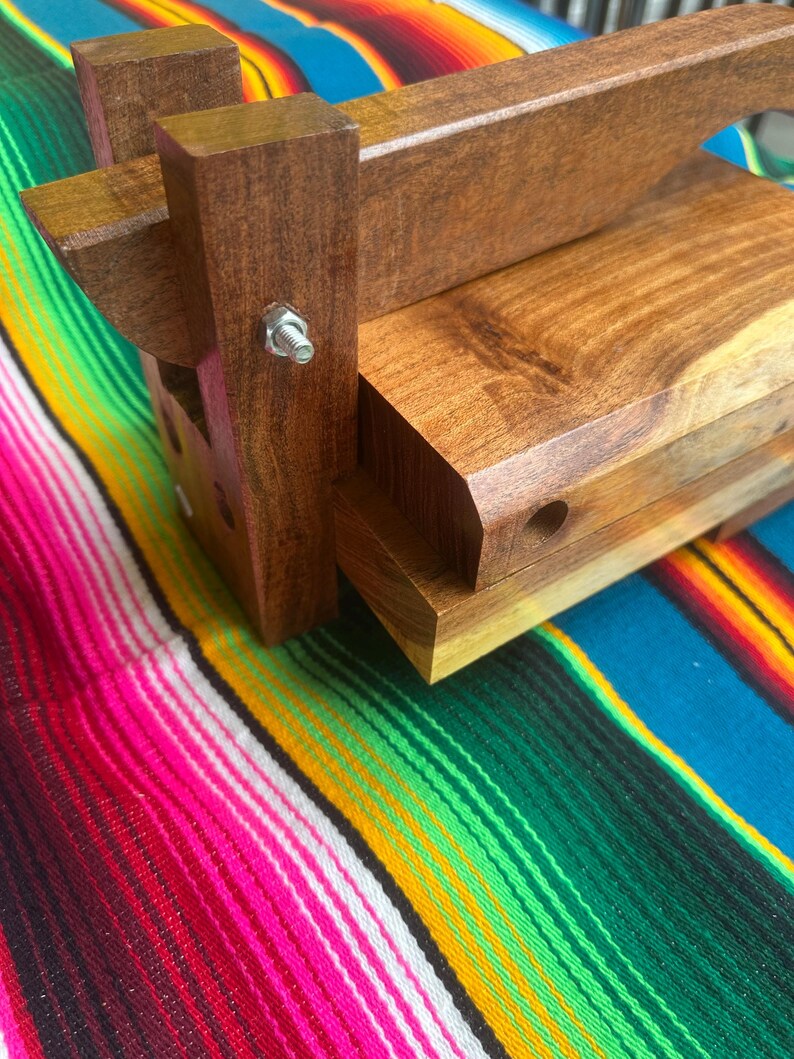 Op de afbeelding: Een houten tortillapers met een donkerbruine afwerking. De pers heeft een rechthoekige basis en een scharnierend bovendeel met een handvat. Een metalen bout zet de componenten van de pers vast. De pers staat tegen een kleurrijke gestreepte textielachtergrond.