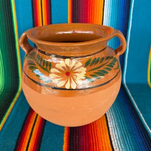 May include: A round terracotta clay pot with two side handles, featuring a glazed upper section decorated with a hand-painted cream-colored flower and green leaves, set against a smooth, unglazed matte terracotta base.