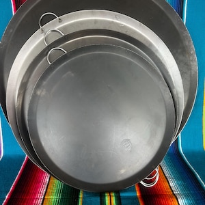 May include: A stack of four round, metal paella pans of varying sizes. The pans are dark gray and silver, with small, curved handles. The pans are arranged on a colorful striped textile background.