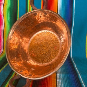May include: A hammered copper bowl with two rounded handles. The bowl has a textured interior and a shiny, reflective surface. The copper has a warm, reddish-brown hue. The bowl is set against a colorful striped textile background.