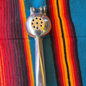 May include: A shiny, silver citrus juicer with a round head featuring multiple holes and a long handle. The juicer is set against a colorful striped background of red, orange, and blue.