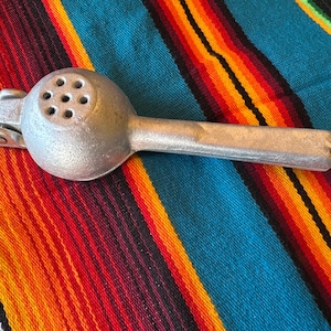 May include: A silver-colored citrus juicer with a round head featuring multiple holes and a long handle. The juicer is made of metal and is resting on a colorful striped textile.