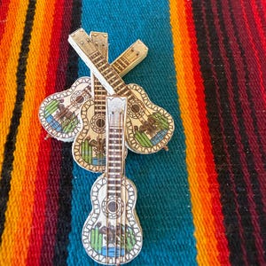 May include: Three small wooden guitar-shaped ornaments featuring a hand-painted design of a brown eagle perched on a green cactus against a blue background. Each guitar has a light tan body with dark brown fretboard details and thin wire strings.