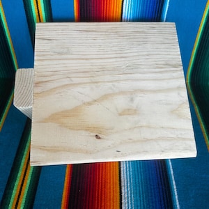 Puede incluir: Un cuadrado de madera sin terminar de color claro con un soporte de madera m&aacute;s peque&ntilde;o adjunto a un lado. La veta de la madera es visible. El cuadrado est&aacute; sobre un fondo a rayas colorido.