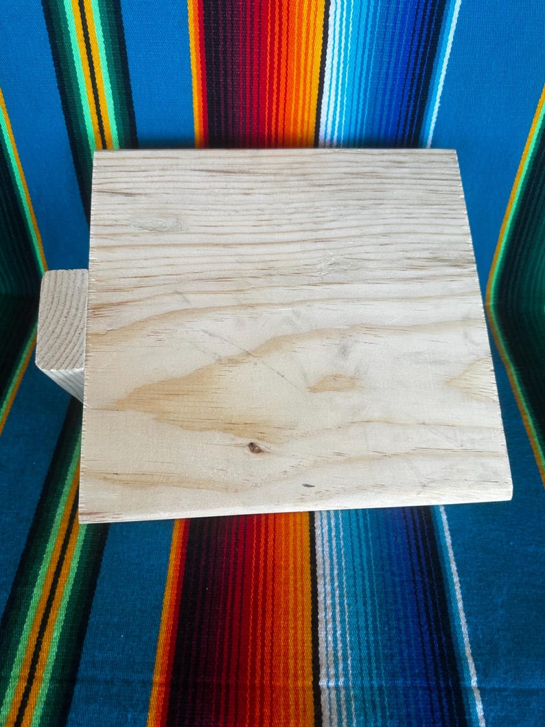 Puede incluir: Un cuadrado de madera sin terminar de color claro con un soporte de madera m&aacute;s peque&ntilde;o adjunto a un lado. La veta de la madera es visible. El cuadrado est&aacute; sobre un fondo a rayas colorido.