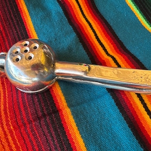 May include: A silver-colored citrus juicer with a round head featuring multiple holes and a long handle. The juicer is resting on a colorful striped textile with shades of red, orange, yellow, and blue.