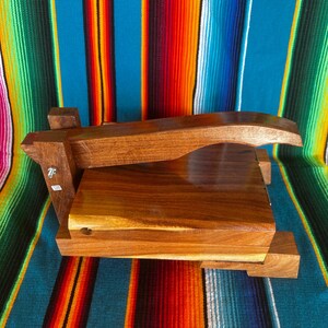 Op de afbeelding: Een handgemaakte houten tortillapers met een rechthoekige basis en een gebogen handvat. De pers is gemaakt van donker hout en staat op een kleurrijke gestreepte textielachtergrond.