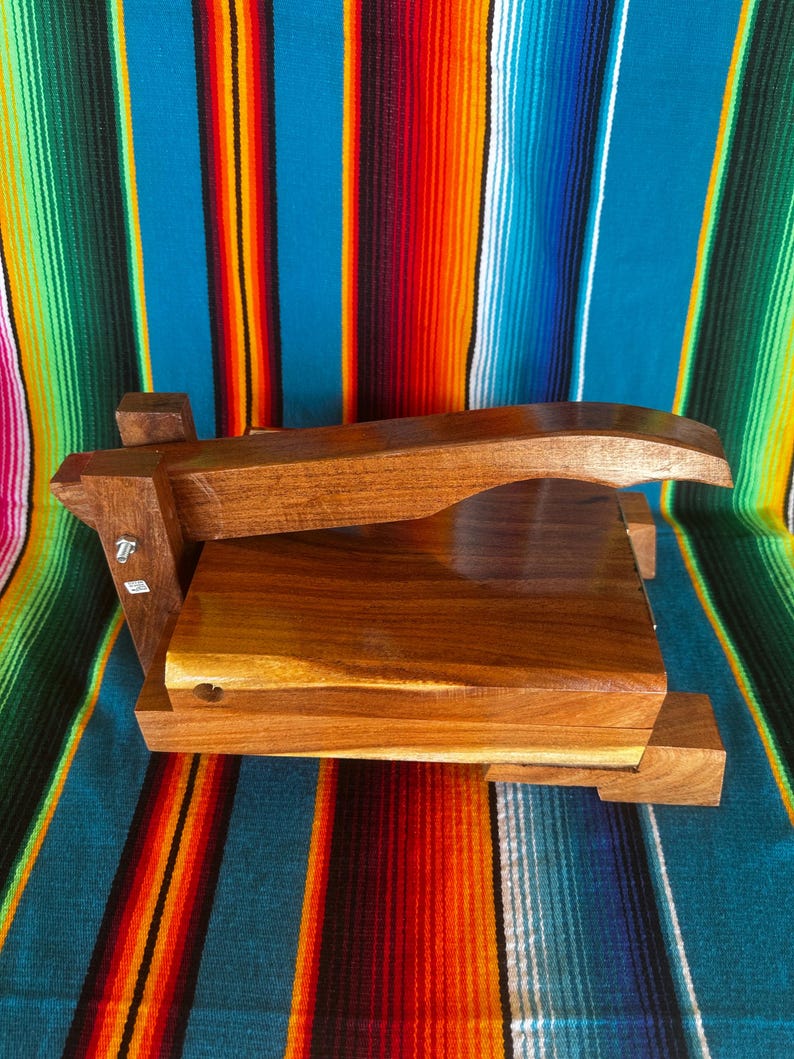 Op de afbeelding: Een handgemaakte houten tortillapers met een rechthoekige basis en een gebogen handvat. De pers is gemaakt van donker hout en staat op een kleurrijke gestreepte textielachtergrond.