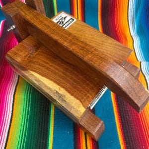 Op de afbeelding: Een houten tortillapers met een rechthoekige basis en een scharnierend deksel. De pers is gemaakt van donkerbruin hout en heeft een handvat om tortilla's te persen. Het label op de pers luidt "Mexico". De pers staat tegen een kleurrijke gestreepte achtergrond.