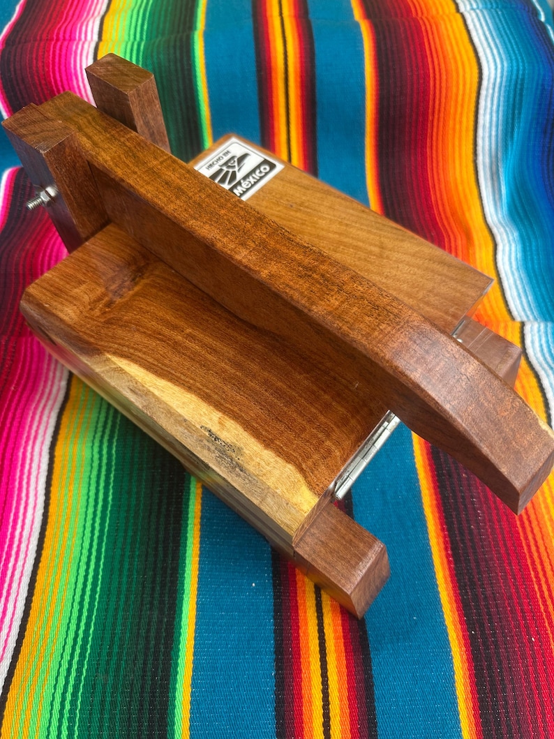 Op de afbeelding: Een houten tortillapers met een rechthoekige basis en een scharnierend deksel. De pers is gemaakt van donkerbruin hout en heeft een handvat om tortilla's te persen. Het label op de pers luidt "Mexico". De pers staat tegen een kleurrijke gestreepte achtergrond.