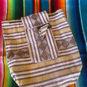 May include: A beige and brown striped woven bag with a matching flap closure. The bag is set against a colorful striped textile background, featuring shades of pink, green, yellow, and blue.