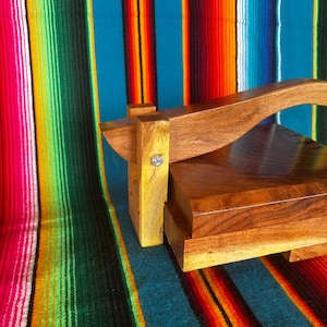 Op de afbeelding: Een houten tortillapers met een gebogen handvat en een rechthoekige basis. De pers is gemaakt van donkerbruin hout en staat tegen een kleurrijke gestreepte achtergrond.