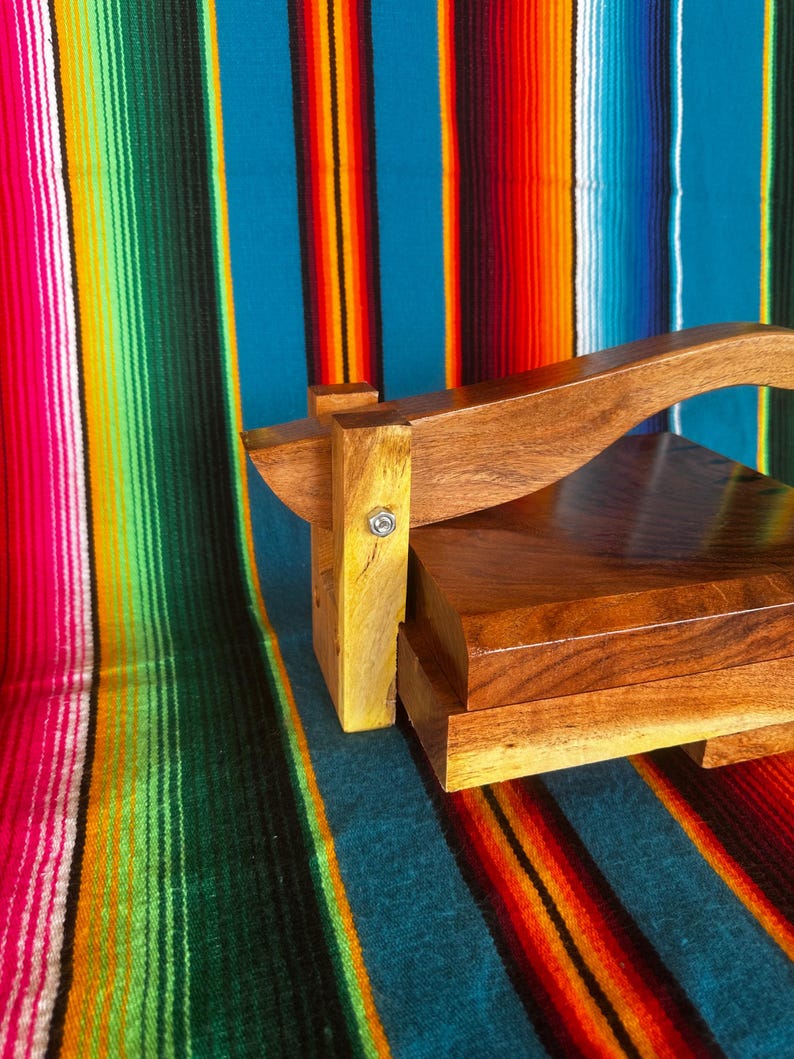 Op de afbeelding: Een houten tortillapers met een gebogen handvat en een rechthoekige basis. De pers is gemaakt van donkerbruin hout en staat tegen een kleurrijke gestreepte achtergrond.
