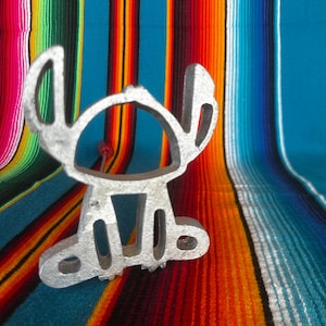May include: A silver-colored metal figure of a cartoon character with large ears and a small red pin. The figure is set against a colorful striped background with shades of blue, orange, red, and yellow.