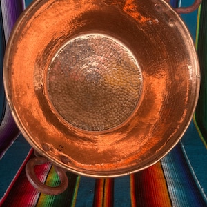 May include: A hammered copper bowl with two rounded handles. The bowl has a shiny, reflective surface and a textured interior. The background features a colorful striped textile, adding a vibrant contrast to the copper.