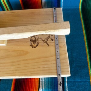Puede incluir: Una prensa de tortillas de madera de color claro con un mango. La prensa mide aproximadamente 30 cm de largo. La imagen incluye una regla y un gr&aacute;fico de una persona montando un toro con la palabra "MEX" al lado.