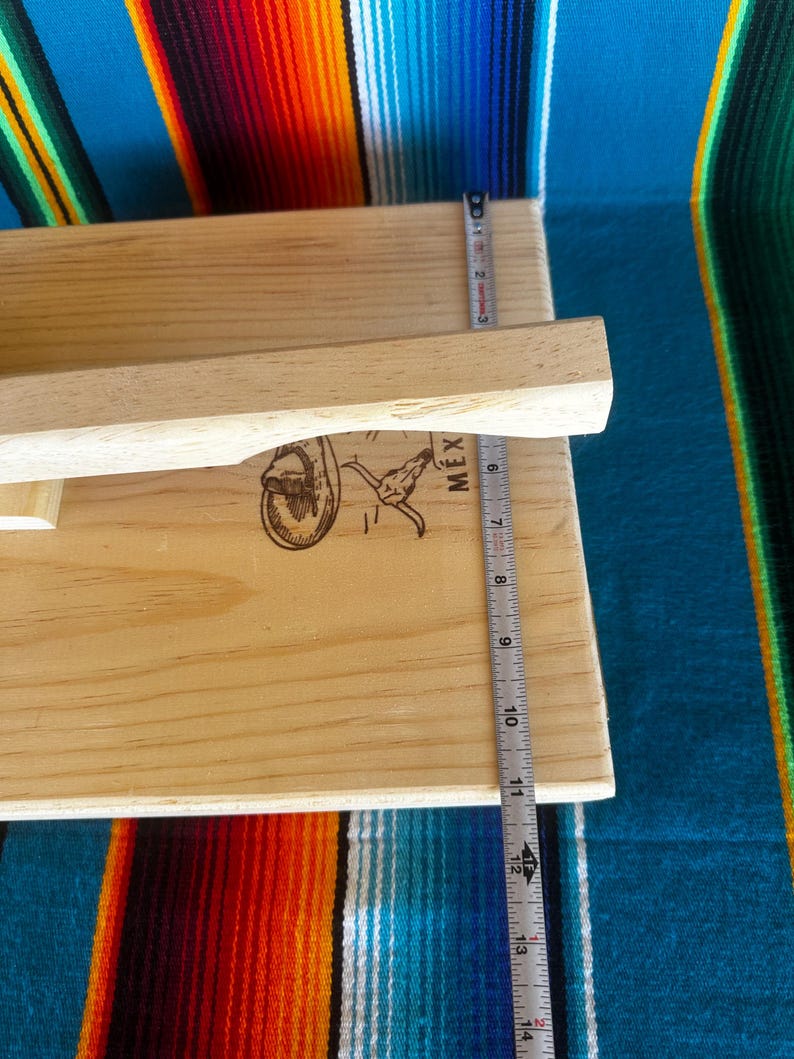 Puede incluir: Una prensa de tortillas de madera de color claro con un mango. La prensa mide aproximadamente 30 cm de largo. La imagen incluye una regla y un gr&aacute;fico de una persona montando un toro con la palabra "MEX" al lado.