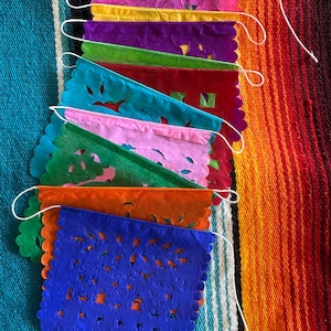May include: A vibrant assortment of colorful papel picado banners. Each banner is a different color, including pink, yellow, purple, green, teal, red, orange, and blue. The banners have intricate cut-out designs and scalloped edges, strung together with white string.