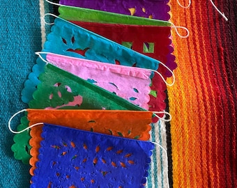 Könnte beinhalten: Eine Auswahl an farbenfrohen Papel Picado-Bannern. Jedes Banner hat eine andere Farbe, darunter Rosa, Gelb, Lila, Grün, Türkis, Rot, Orange und Blau. Die Banner haben aufwendige Ausschnitte und einen gewellten Rand, die mit weißer Schnur verbunden sind.