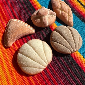 May include: An assortment of Mexican sweet bread, including conchas, empanadas, and other pastries, arranged on a colorful striped textile. The baked goods are light brown and dusted with sugar, showcasing various shapes and textures.