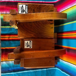 Op de afbeelding: Houten boekenplank met een gebogen plankontwerp. De unit is gemaakt van donkerbruin hout en heeft de tekst "Hecho en M&eacute;xico" op een sticker. De unit wordt weergegeven op een kleurrijk gestreept textiel.