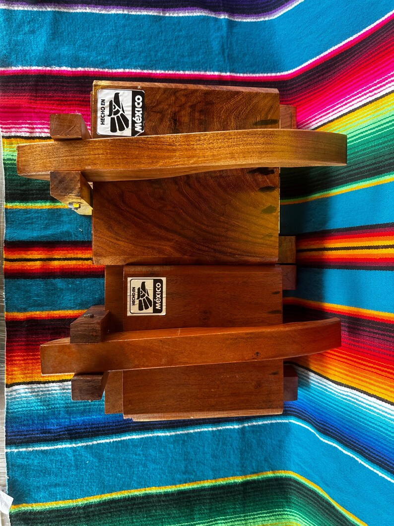 Op de afbeelding: Houten boekenplank met een gebogen plankontwerp. De unit is gemaakt van donkerbruin hout en heeft de tekst "Hecho en M&eacute;xico" op een sticker. De unit wordt weergegeven op een kleurrijk gestreept textiel.
