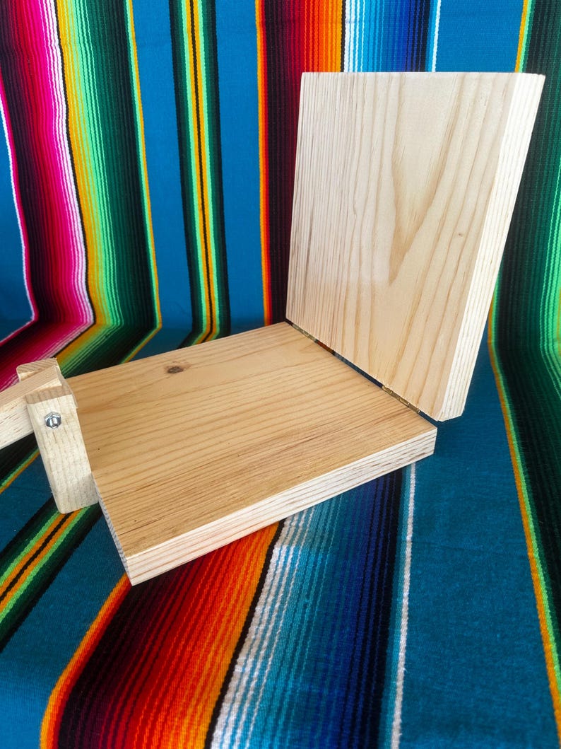 Puede incluir: Una prensa de tortillas de madera clara, con una base rectangular y una tapa con bisagras. La prensa est&aacute; hecha de madera sin terminar y est&aacute; dise&ntilde;ada para hacer tortillas. El fondo es un textil a rayas de colores.