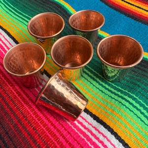 May include: Six copper shot glasses with a hammered texture are arranged on a colorful striped textile. The shot glasses are cylindrical with a flared rim and a shiny, reflective surface. The textile features stripes of red, green, blue, and yellow.