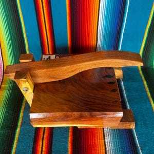 Op de afbeelding: Een houten tortillapers met een gebogen handvat en een rechthoekige basis. De pers is gemaakt van donkerbruin hout en staat op een kleurrijke gestreepte deken met turquoise, oranje en groene strepen.