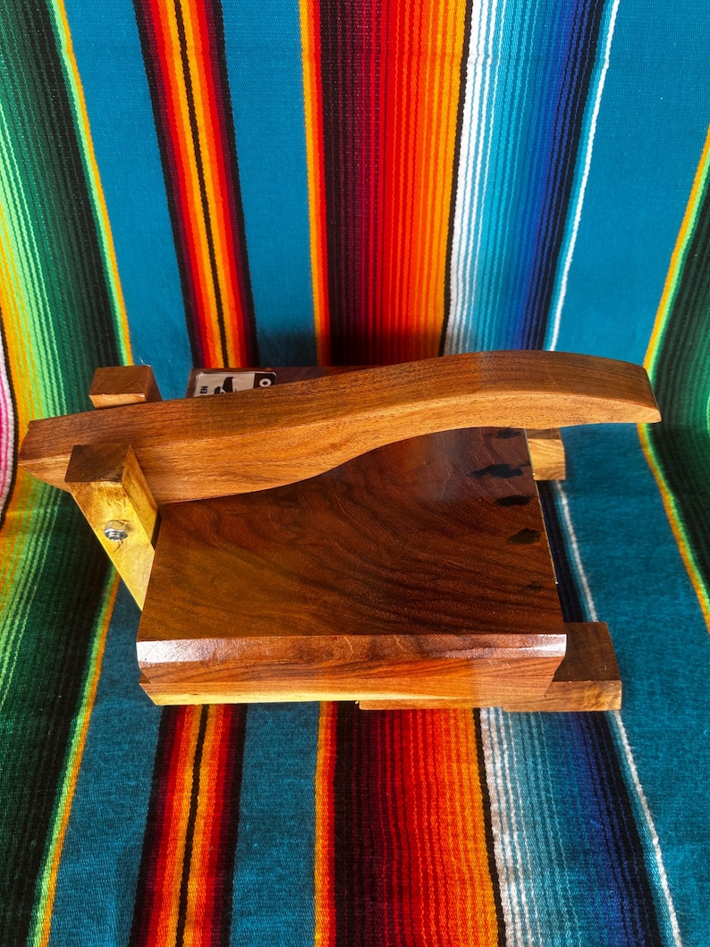 Op de afbeelding: Een houten tortillapers met een gebogen handvat en een rechthoekige basis. De pers is gemaakt van donkerbruin hout en staat op een kleurrijke gestreepte deken met turquoise, oranje en groene strepen.