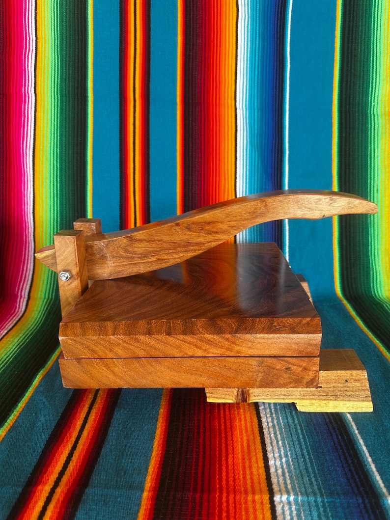 Op de afbeelding: Een houten tortillapers met een lange handgreep en een rechthoekige basis. De pers is gemaakt van lichtbruin hout en heeft een eenvoudig, functioneel ontwerp. Het is ontworpen om tortilla's te maken. De achtergrond is een kleurrijk gestreept textiel.