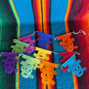 May include: A decorative garland featuring multiple small, die-cut paper skulls in vibrant colors including purple, green, yellow, orange, blue, and red. Each skull is suspended from a thin white string, creating a festive, multi-colored banner.