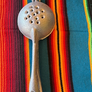 May include: A silver-colored, cast-iron kitchen tool with a round, perforated head and a long handle. The tool has a loop at the top and is designed for straining or skimming. The background is a colorful striped textile.