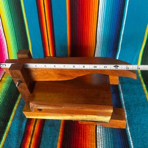 Op de afbeelding: Een houten tortillapers met een rechthoekige basis en een scharnierende bovenkant. De pers is gemaakt van donkerbruin hout en meet ongeveer 33 cm in lengte. Het rust op een kleurrijk gestreept textiel.