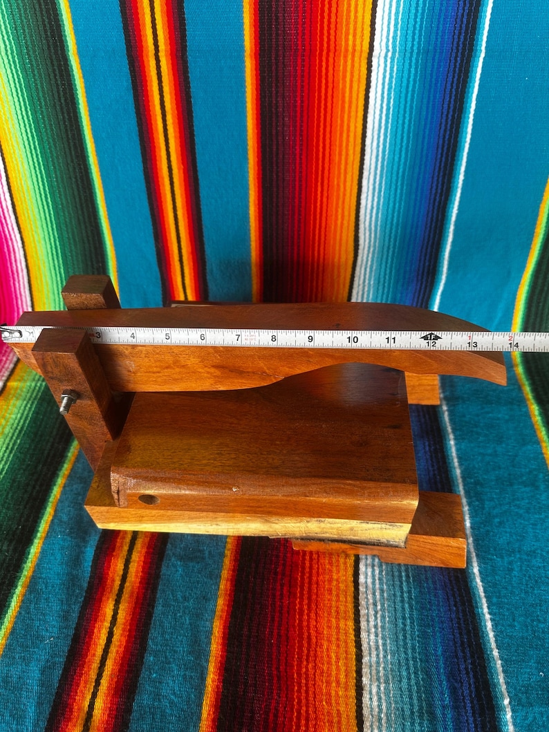 Op de afbeelding: Een houten tortillapers met een rechthoekige basis en een scharnierende bovenkant. De pers is gemaakt van donkerbruin hout en meet ongeveer 33 cm in lengte. Het rust op een kleurrijk gestreept textiel.