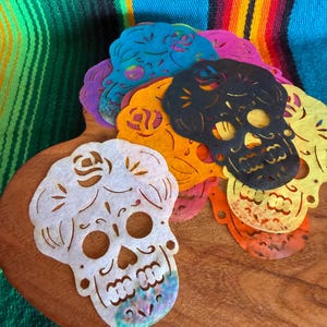 May include: A collection of die-cut sugar skull shapes made from thin, colorful material. The skulls feature intricate floral and geometric cutouts, including eye sockets, nasal cavities, and teeth. Colors include white, black, orange, yellow, and blue.