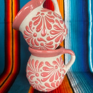 May include: Two stacked, handmade ceramic mugs. The mugs are white with a pink floral design and pink trim. Each mug has a handle. The background is a colorful striped textile.