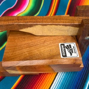 Op de afbeelding: Een houten tortillapers met een rechthoekige basis en een scharnierende bovenkant. De pers is gemaakt van licht en donkerbruin hout en heeft een sticker met "Mexico" en "Hecho en".