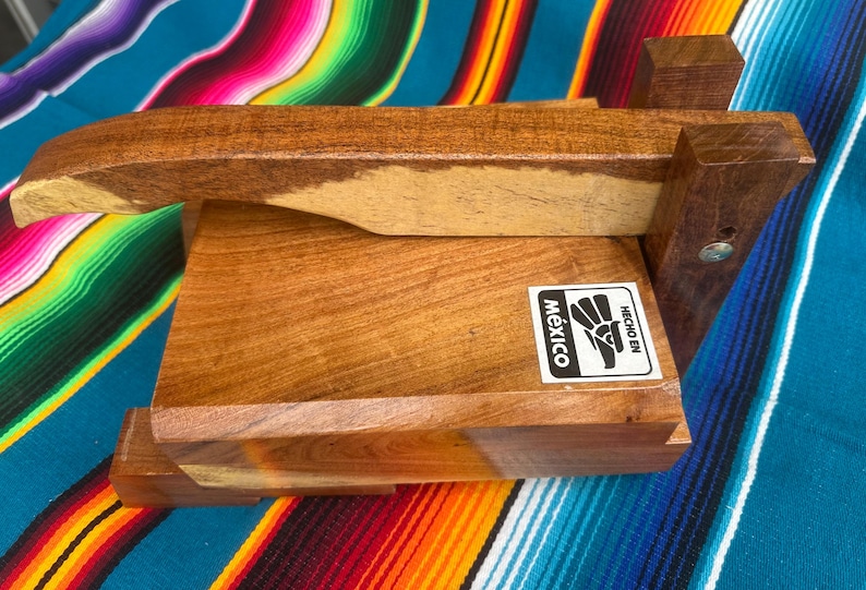 Op de afbeelding: Een houten tortillapers met een rechthoekige basis en een scharnierende bovenkant. De pers is gemaakt van licht en donkerbruin hout en heeft een sticker met "Mexico" en "Hecho en".