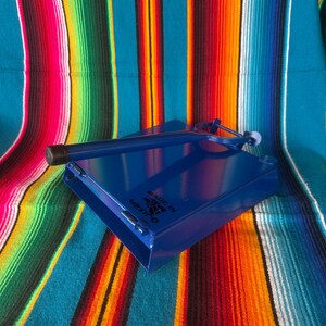 K&ouml;nnte beinhalten: Blaue Tortillapresse aus Metall mit Griff und kleinem wei&szlig;en Ball am Ende. Die Presse liegt auf einer bunten gestreiften Decke mit roten, orangefarbenen, gelben und blauen Streifen. Der Text "MADE IN MEXICO" ist sichtbar.