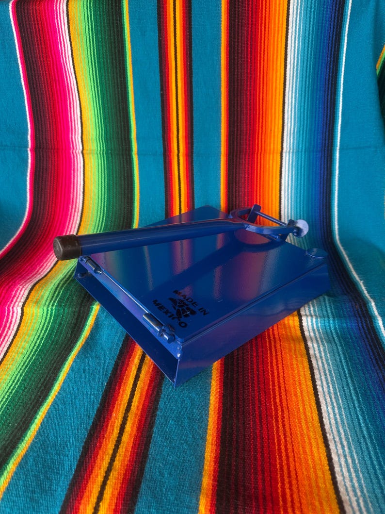 K&ouml;nnte beinhalten: Blaue Tortillapresse aus Metall mit Griff und kleinem wei&szlig;en Ball am Ende. Die Presse liegt auf einer bunten gestreiften Decke mit roten, orangefarbenen, gelben und blauen Streifen. Der Text "MADE IN MEXICO" ist sichtbar.