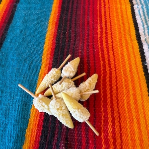 May include: A cluster of small, pale yellow, cone-shaped food items, each pierced with a thin wooden skewer. The items have a textured, granular surface and are arranged in a loose pile, showing their pointed ends and wooden stick handles.