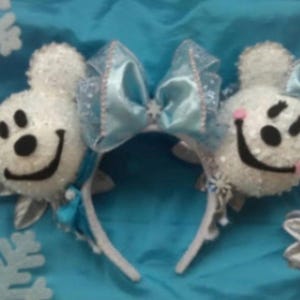 May include: A pair of white, textured mouse-ear headbands featuring a large, light blue, translucent bow with silver accents. Each ear is decorated with a smiling face, black eyes, a black nose, and a wide black mouth, with pink cheeks on one side.