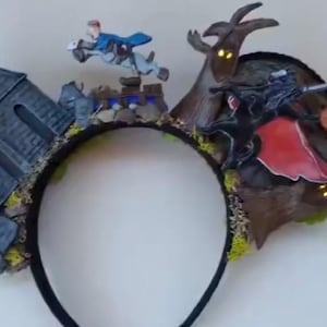 May include: A headband featuring a Sleepy Hollow theme with a dark gray church, a figure on a horse, a headless horseman with a red cape, glowing orange pumpkin lights, and brown trees with small yellow illuminated eyes set against a black band.