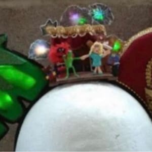 Puede incluir: Un tocado caprichoso con un diseño de hoja verde con acentos brillantes, un escenario en miniatura con figuras y un elemento circular rojo con un borde dorado. El tocado está adornado con pequeñas luces brillantes.