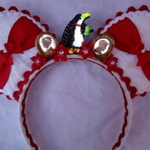 May include: A whimsical headband featuring large, round ears with red and white accents. A black and white penguin figurine sits atop the headband, flanked by cameo-style embellishments and red floral accents. The headband is trimmed with a scalloped edge.