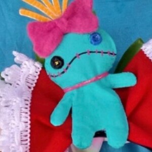 K&ouml;nnte beinhalten: Eine t&uuml;rkisfarbene Pl&uuml;schpuppe mit einer leuchtend pinken Filzschleife, gelben Stachelhaaren und einem aufgestickten Mund. Die Puppe hat ein schwarzes und ein blaues Knopfauge und liegt auf einem roten Stoffhintergrund mit wei&szlig;er Spitzenborte.