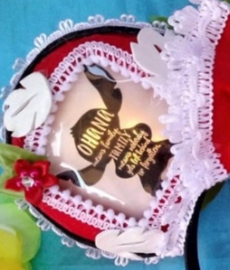 K&ouml;nnte beinhalten: Ein rotes Stirnband mit wei&szlig;er Spitzenborte, wei&szlig;en blattf&ouml;rmigen Akzenten und einer kleinen roten Blume mit einem zentralen Kristall. In der Mitte befindet sich eine kreisf&ouml;rmige Grafik mit dem Text OHANA means family, means nobody gets left behind or forgotten.