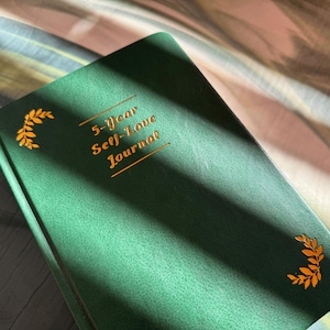 May include: A green 5-Year Self-Love Journal with gold lettering and leaf accents. The journal is closed and has a textured cover. The title is centered on the cover.