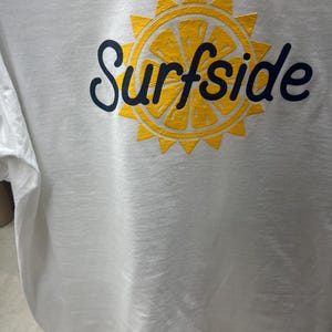 May include: White t-shirt with a yellow and navy blue graphic. The graphic features a yellow citrus fruit slice with a sunburst design and the word "Surfside" in navy blue script.